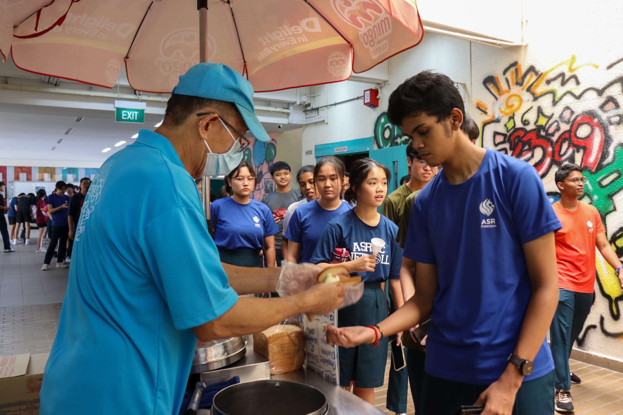 A masked vendor in a blue cap serves food to a line of students in blue shirts at a school canteen stall.